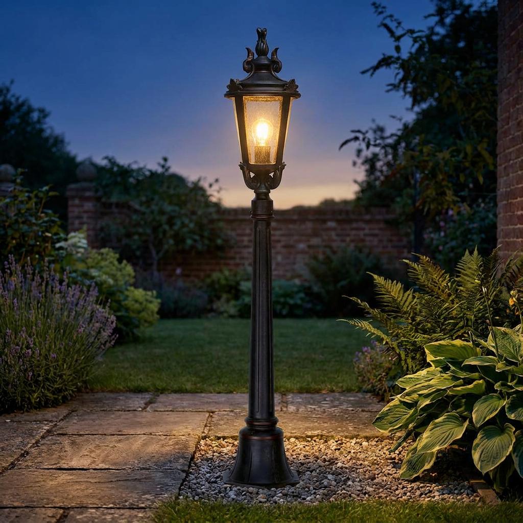 Traditional weathered bronze bollard light with a glowing bulb illuminating a garden path at dusk.