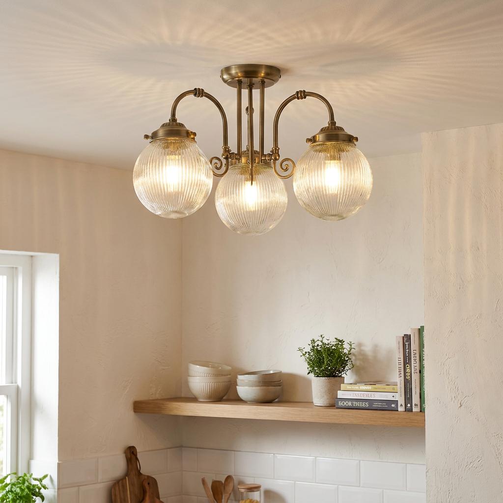 Three-light antique brass flush ceiling fixture with ribbed glass globes illuminated in a neutral-toned kitchen setting.