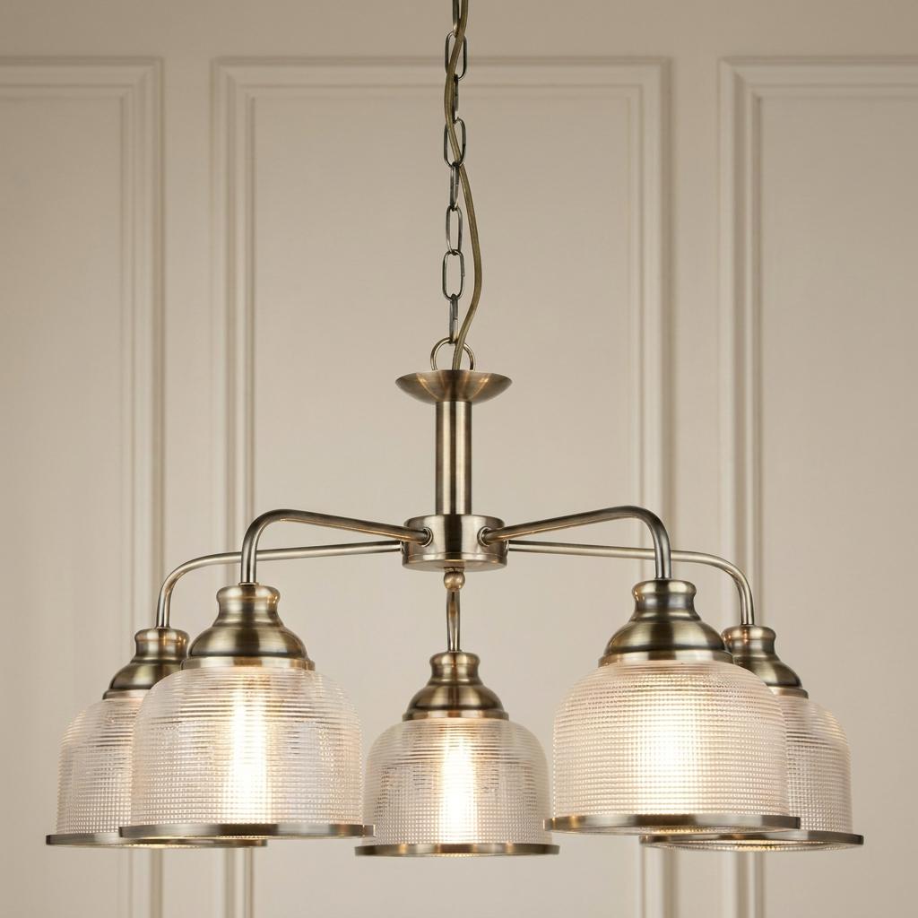 Bistro II five-light antique brass pendant chandelier with holophane glass shades against a neutral paneled wall.