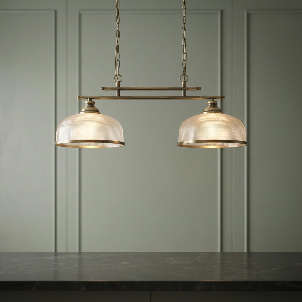 Brushed brass two-light kitchen island fixture with holophane glass shades hanging above a dark countertop.