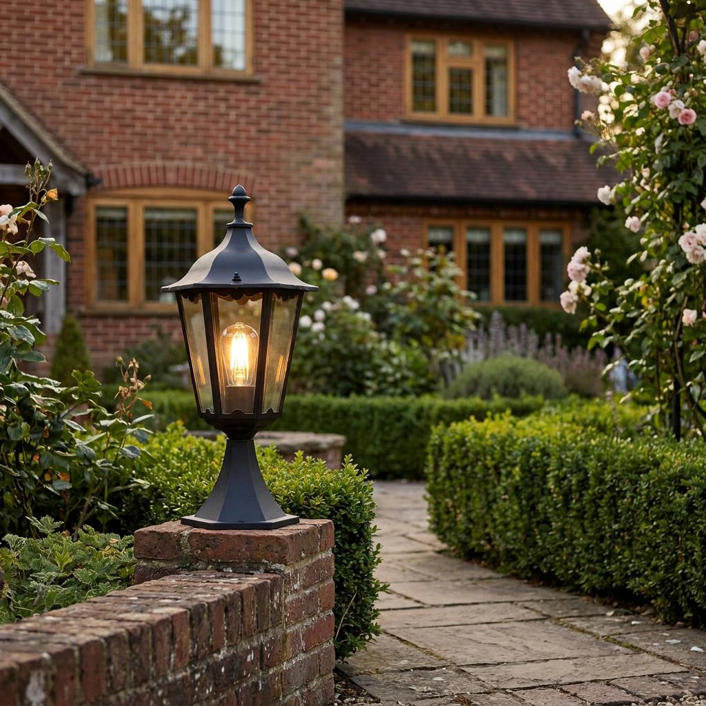 Black lantern outdoor post light glows on a brick pillar in a lush garden setting with a path and brick house.