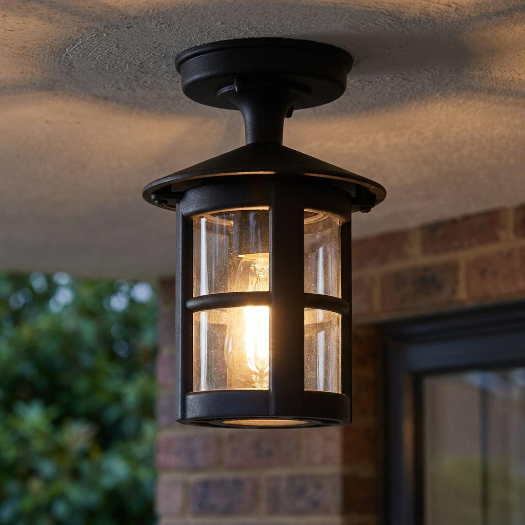 A black metal lantern-style outdoor ceiling light with a glowing vintage bulb mounted on a porch ceiling.