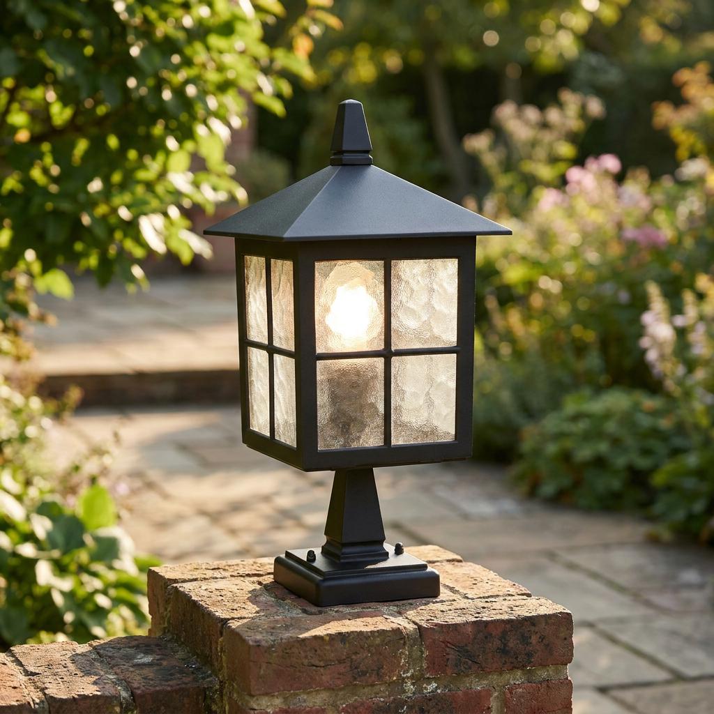 Black outdoor lantern pedestal light with frosted glass panes illuminated on a brick garden wall.