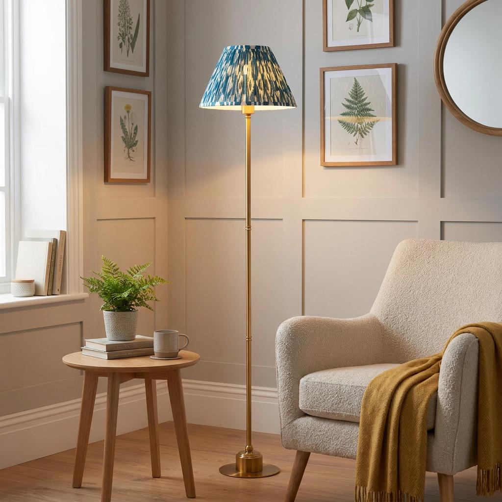 Burley brass floor lamp with blue Ikat shade lit in a living room next to a cream armchair and wooden side table.
