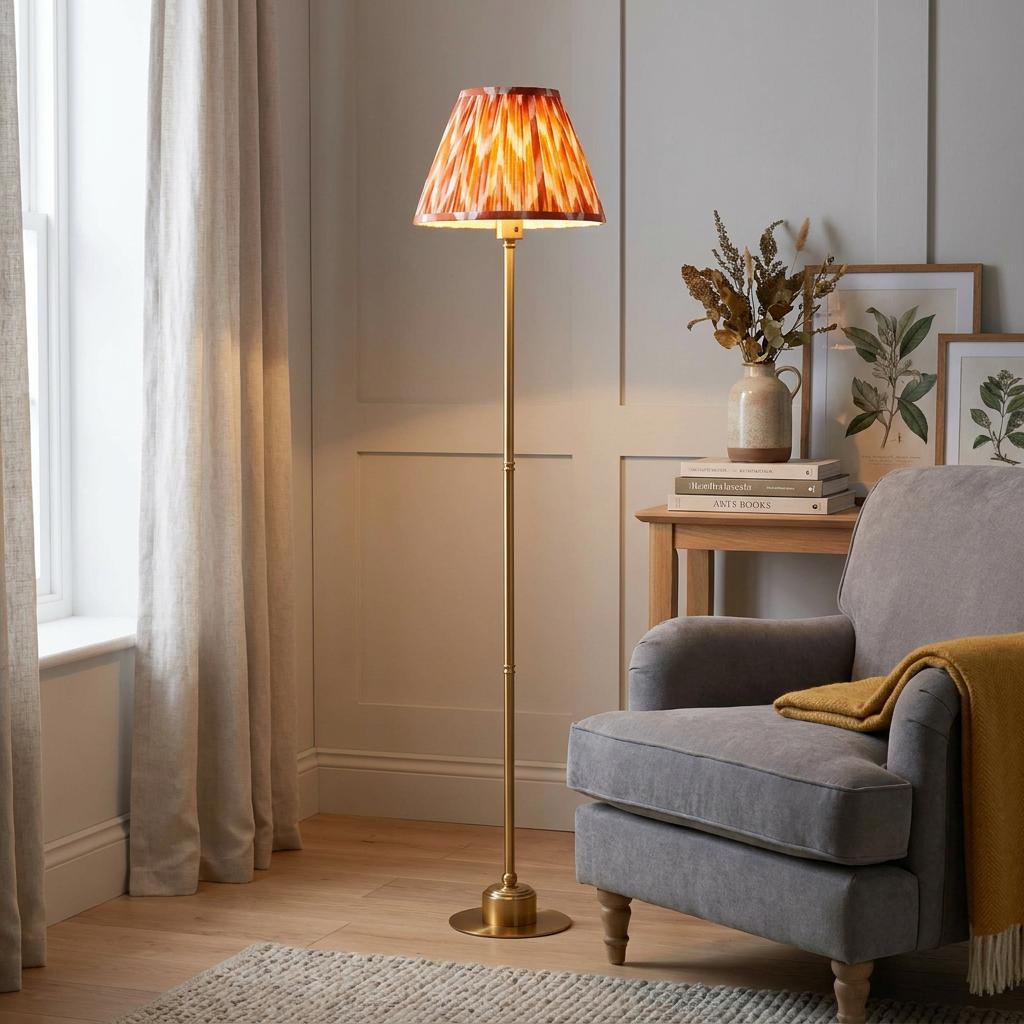 A Burley brass floor lamp with an apricot zigzag shade stands beside a grey armchair in a cozy living room.