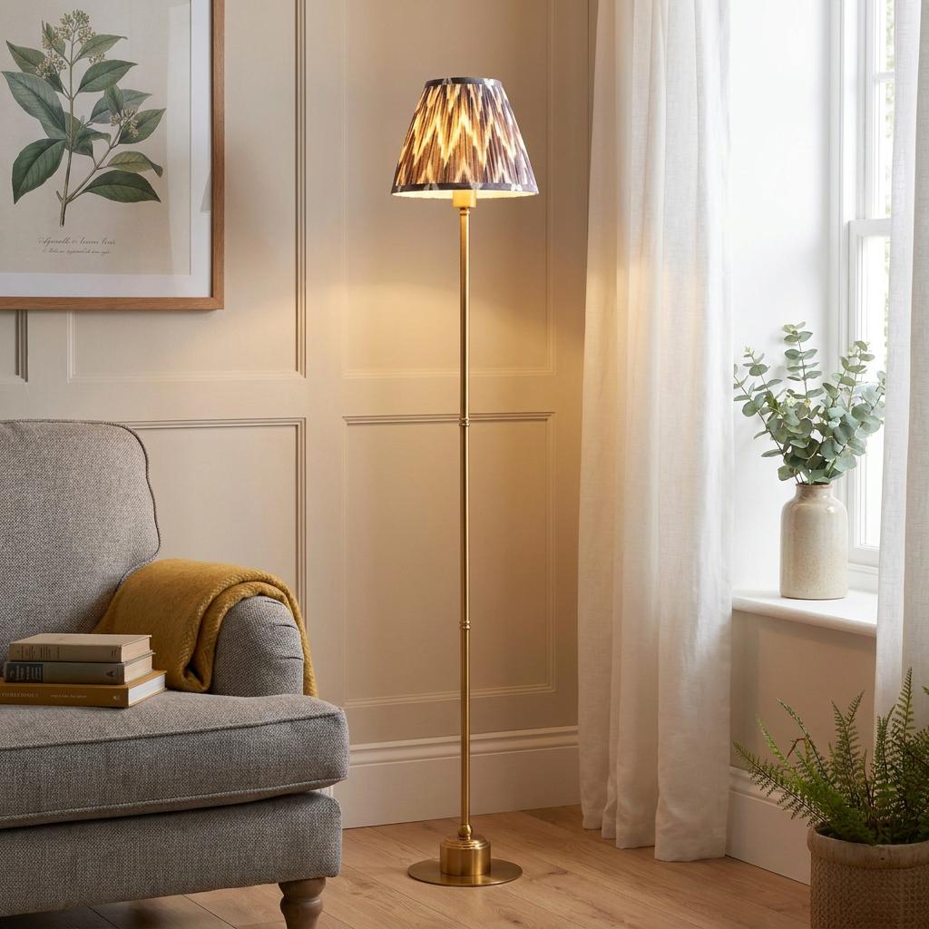Brass floor lamp with a zigzag patterned shade illuminating a cozy living room corner next to a grey armchair.