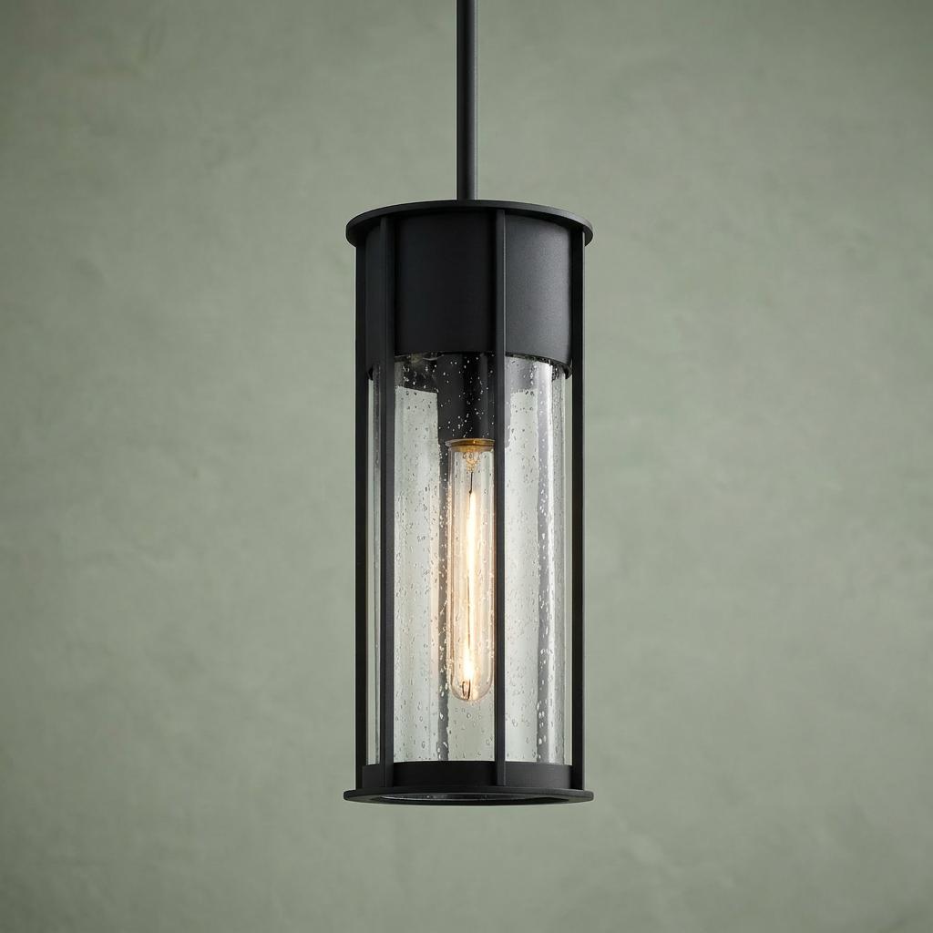 Cylindrical black outdoor pendant light with seeded glass and a warm Edison bulb against a green wall.