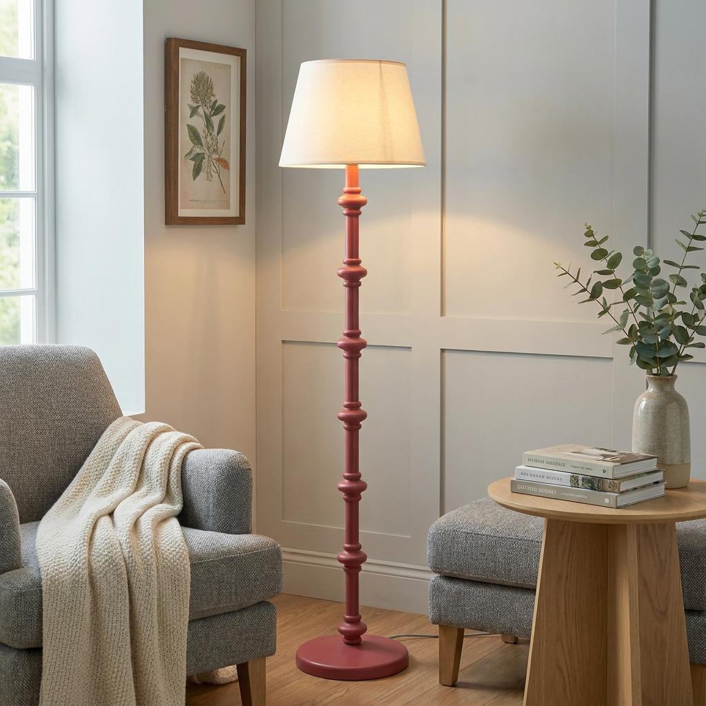 Pink wooden floor lamp with a white drum shade in a living room between a grey armchair and a wooden side table.