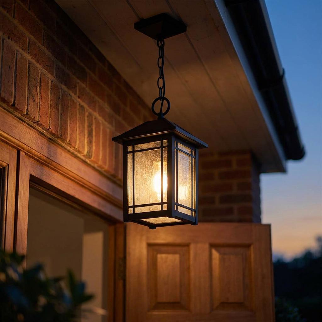A black outdoor chain lantern with seeded glass hanging from an eave, illuminated against a brick wall at dusk.