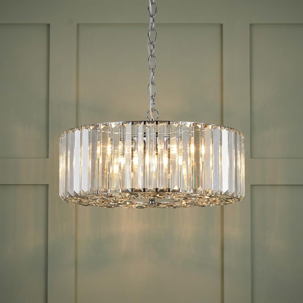 Drum-shaped chrome pendant light with vertical glass crystals glowing against a pale green paneled wall.