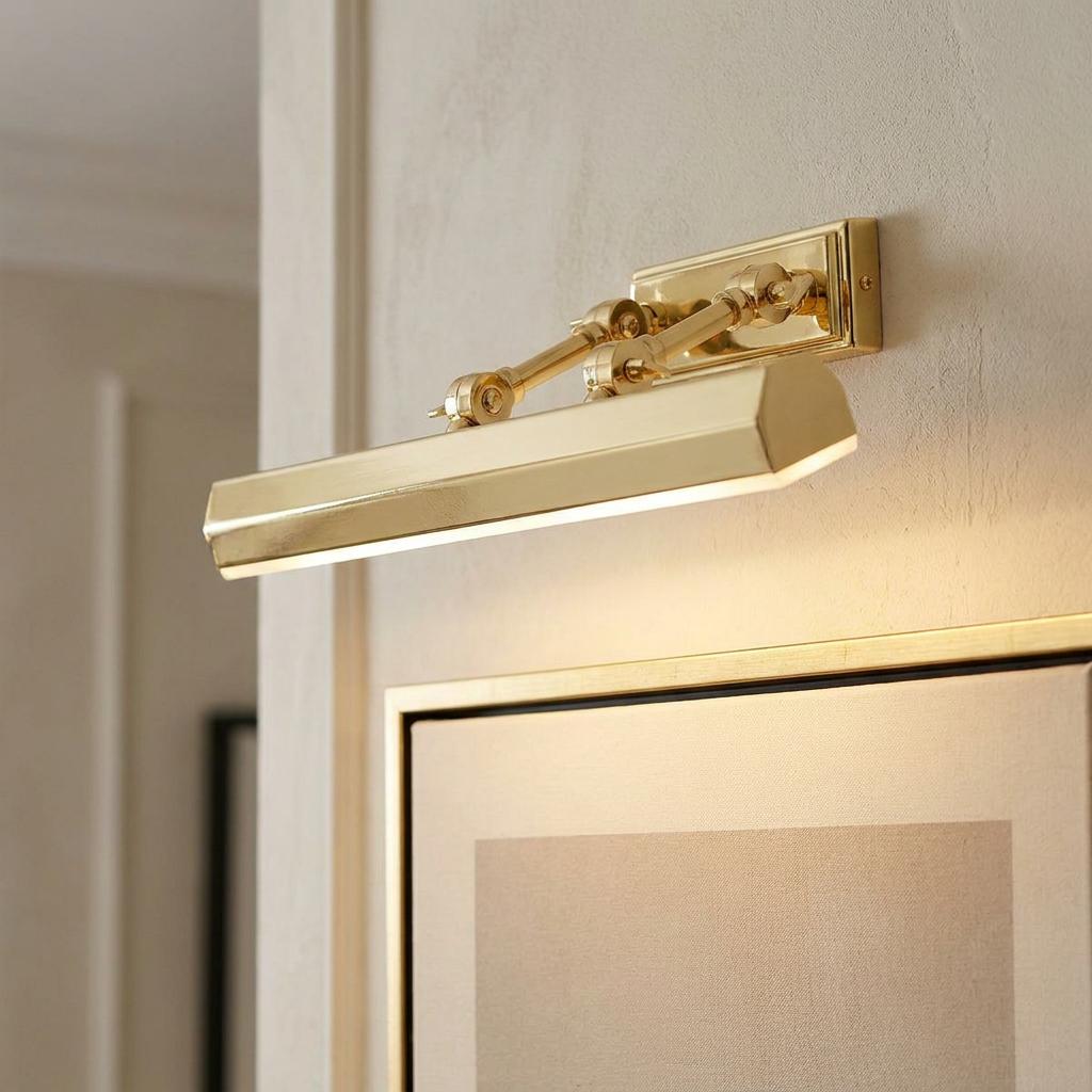 Polished brass picture light with a hexagonal shade mounted on a beige wall above a framed artwork, illuminated.