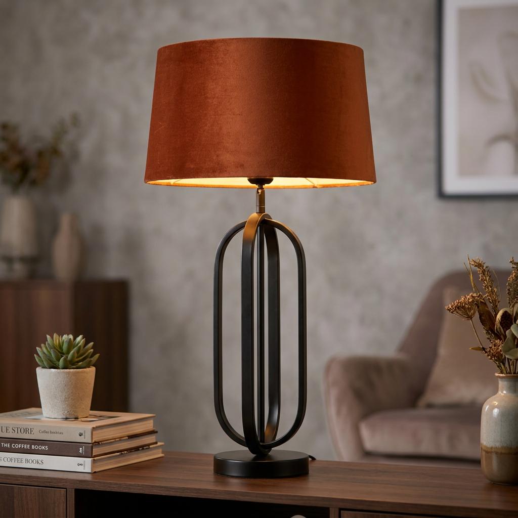 Chic table lamp with a matt black open-frame oval base and an orange velvet drum shade on a wooden table.