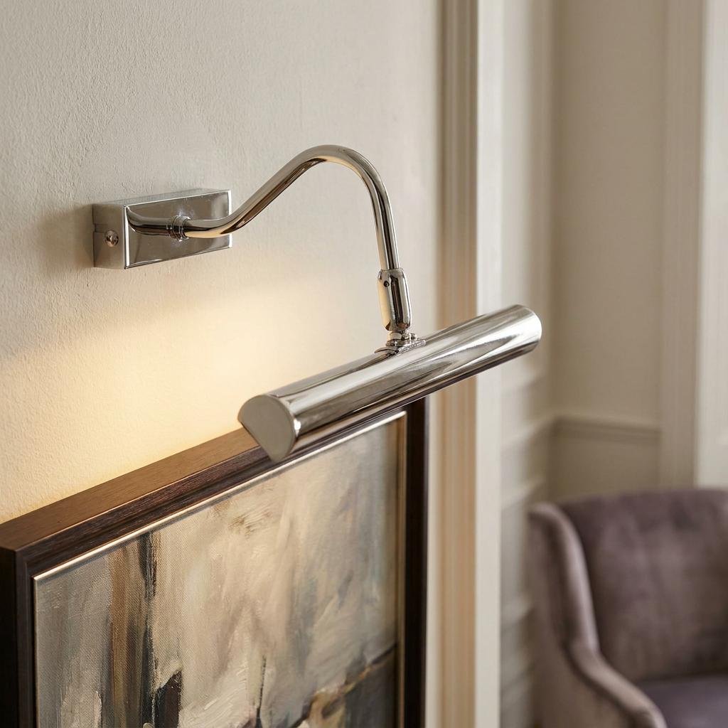 Polished chrome picture light with a curved arm illuminating a framed painting on a neutral wall.