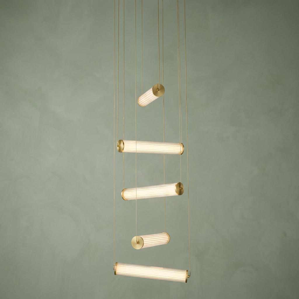 A multi-drop pendant with five horizontal fluted glass cylinders and gold accents hanging against a sage green wall.