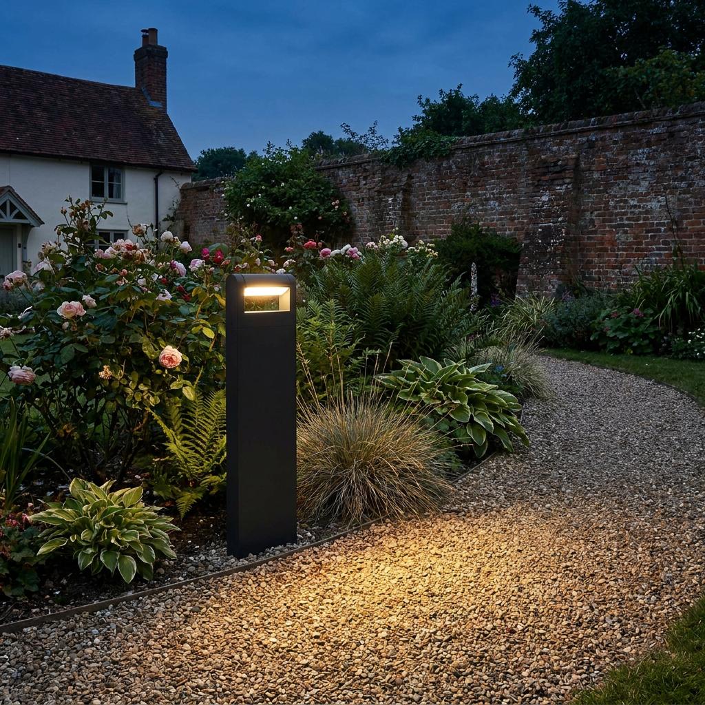 Modern black bollard light illuminating a gravel garden path next to flowering plants at dusk.