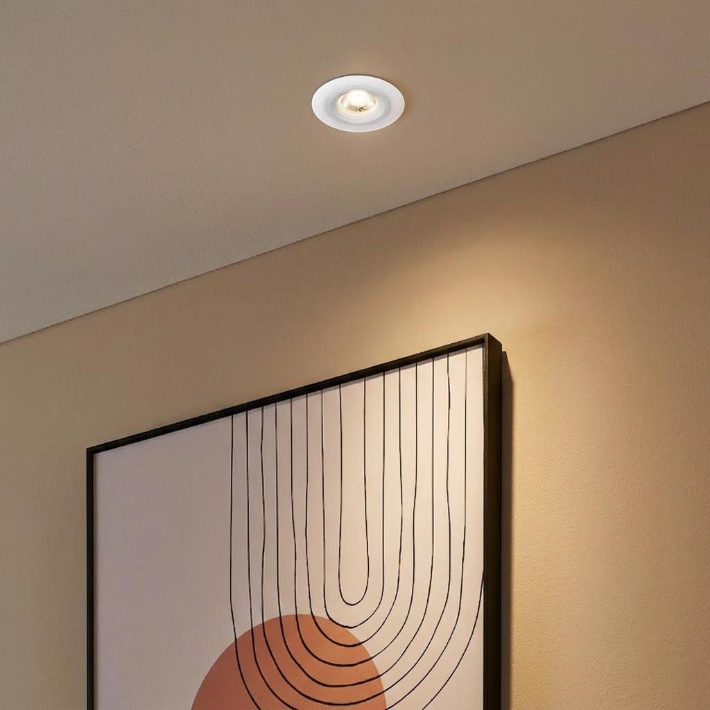 White circular recessed LED spotlight illuminating abstract framed wall art from a neutral ceiling.
