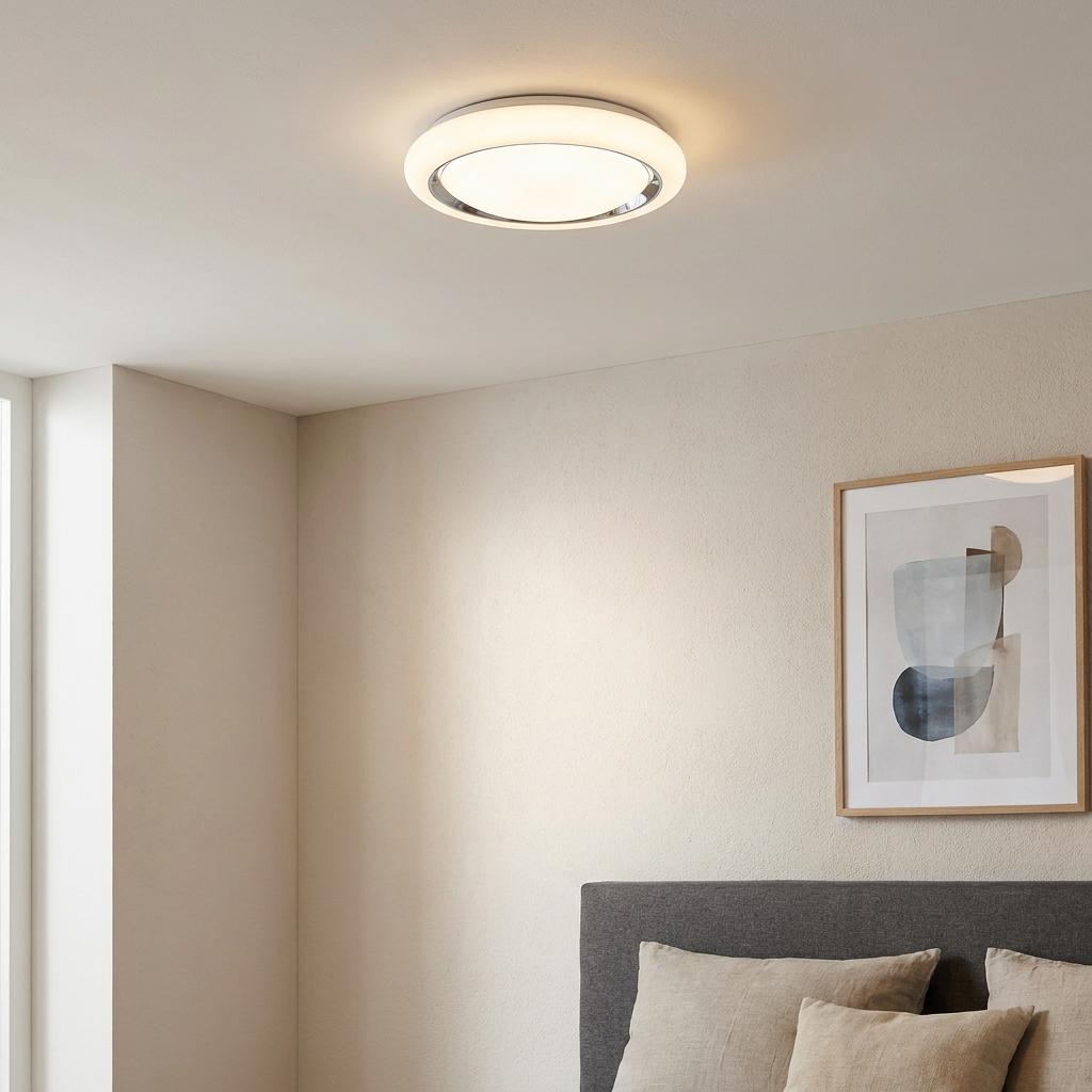 Round white LED flush ceiling light illuminated in a modern bedroom with a grey headboard and framed artwork.