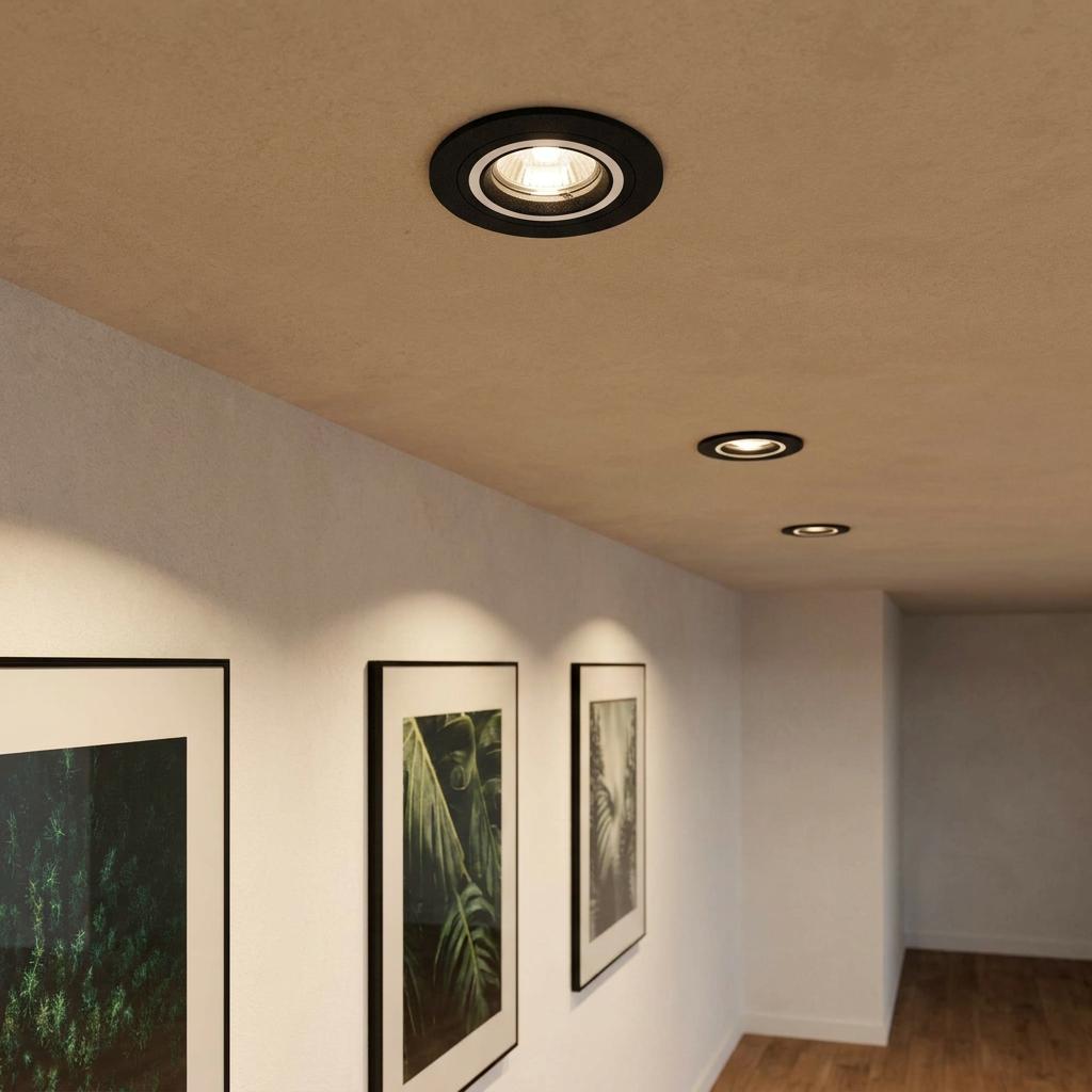 Three black and white recessed spot lights illuminate framed botanical artwork on a white wall in a hallway.
