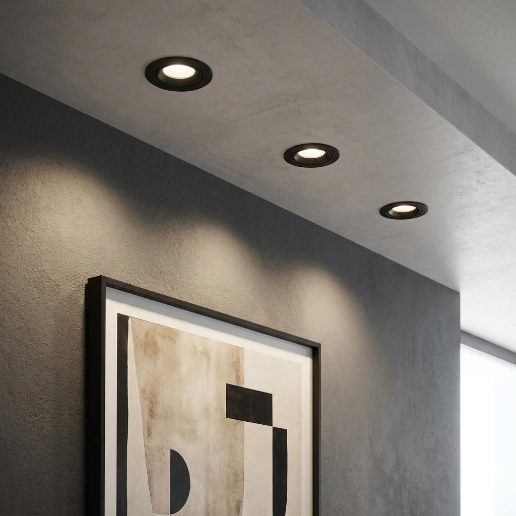 Three black recessed spot lights on a concrete ceiling casting light onto a framed artwork in a modern room.