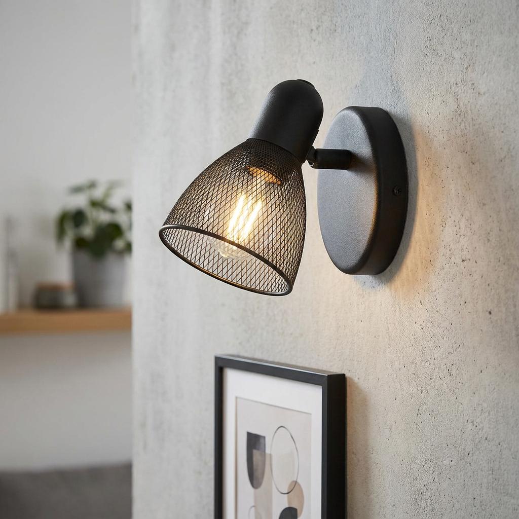 Black mesh wall spotlight with a warm glowing filament bulb mounted on a textured gray wall.