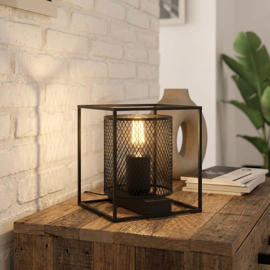 Industrial black metal table lamp with a cube frame and mesh shade on a wooden sideboard against a white brick wall.