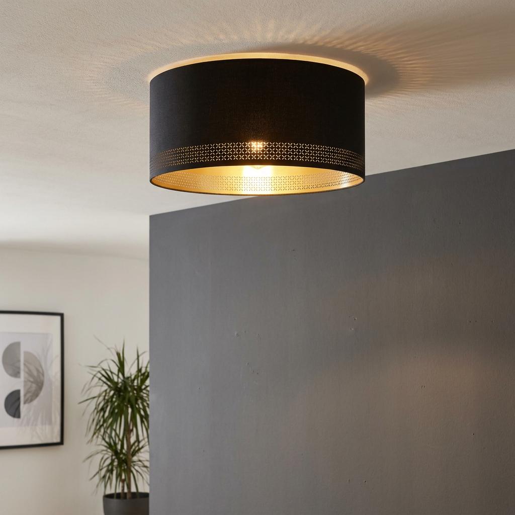 Black drum ceiling light with a gold interior and perforated pattern, mounted on a white ceiling above a grey wall.
