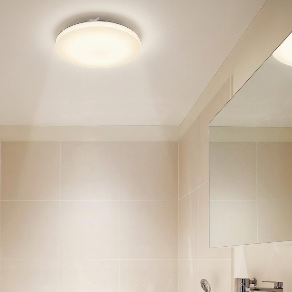 A round, white LED flush ceiling light illuminating a modern bathroom with beige wall tiles.