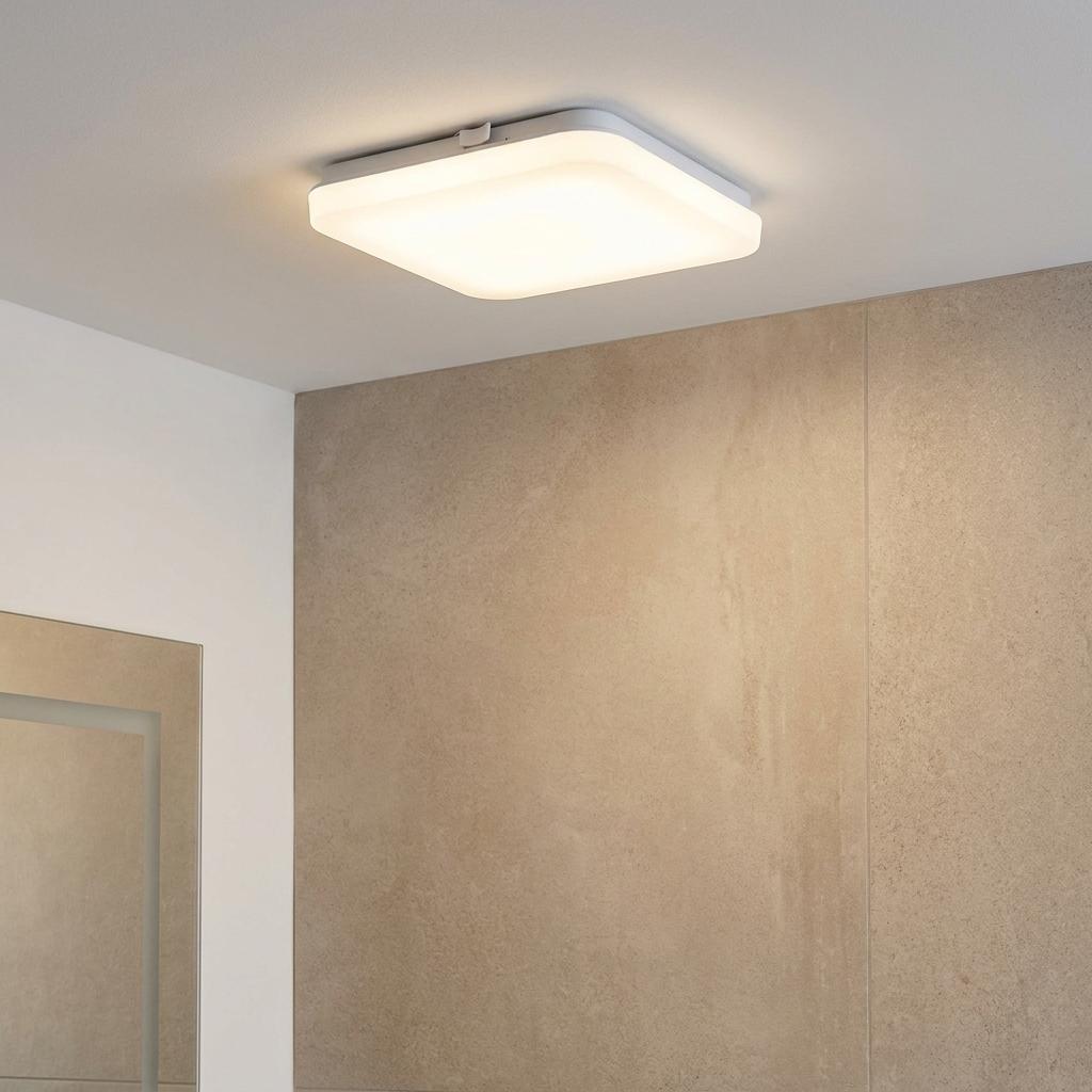 Square white LED flush ceiling light mounted on a bathroom ceiling, emitting a warm glow near a tiled wall.