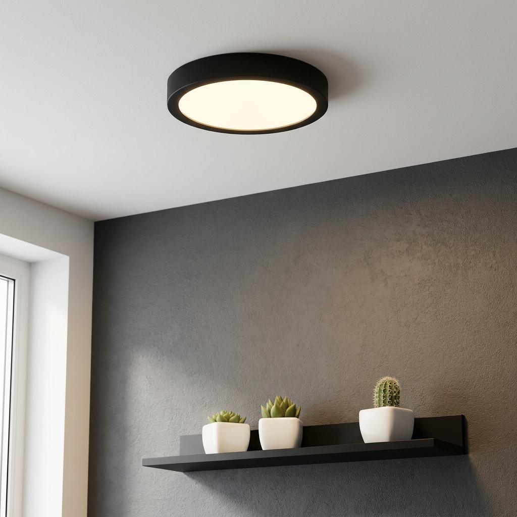 Round black flush-mount LED ceiling light illuminated on a white ceiling above a grey wall with a shelf.