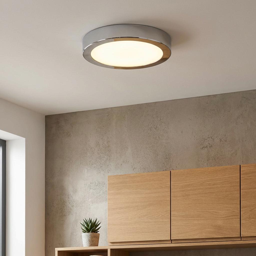 Round chrome flush ceiling light illuminated above modern wooden cabinetry and a grey textured wall.
