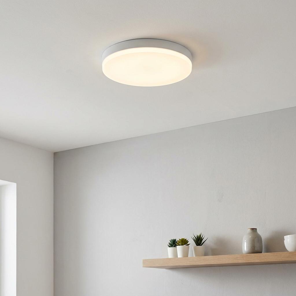 Round white LED flush ceiling light glowing on a white ceiling above a wooden shelf with small plants.