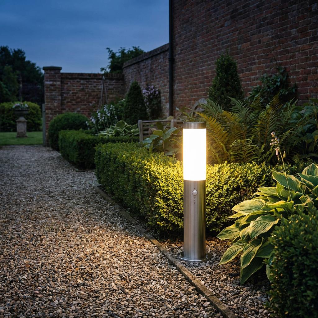 An illuminated stainless steel bollard light with a motion sensor along a gravel path in a garden at dusk.