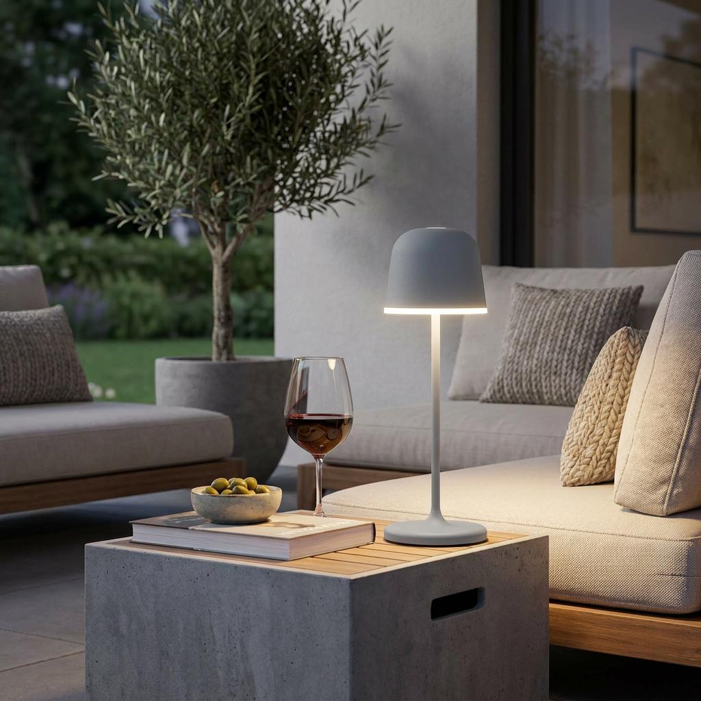 Grey cordless LED outdoor table lamp glowing on a patio table next to a glass of wine and snacks.