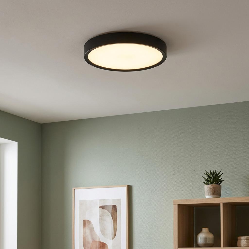 Round black LED flush ceiling light illuminated on a white ceiling in a room with soft green walls.