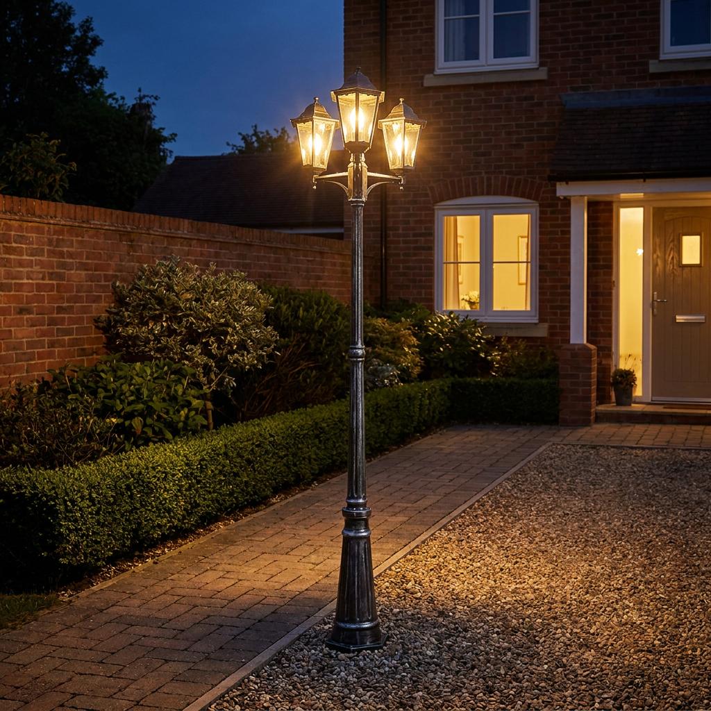 Traditional three-light lamp post illuminating a garden path and driveway outside a brick house at night.