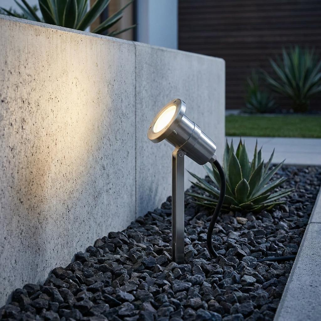 Silver ground spike spotlight illuminating a concrete garden wall from a gravel bed.