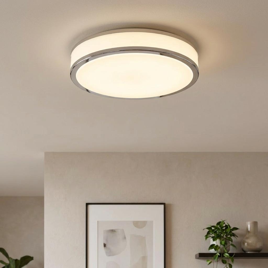 Round chrome LED flush ceiling light illuminating a minimalist room with a picture frame and indoor plant.