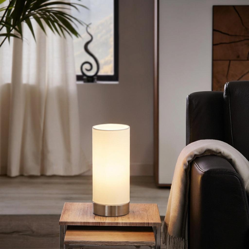 Cylindrical white fabric table lamp with a nickel base glowing on a wooden side table next to a leather chair.