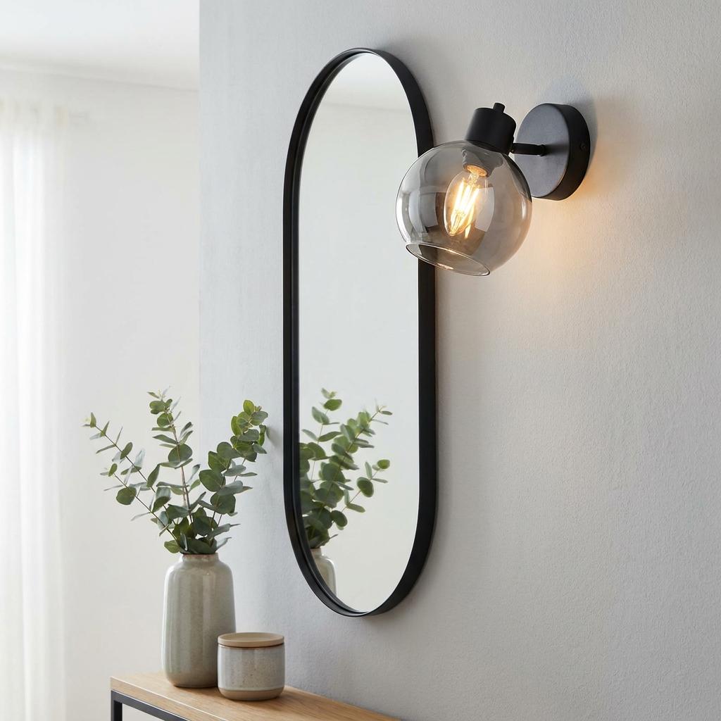 A black wall spotlight with a smoked glass globe shade mounted next to an oval mirror on a light grey wall.