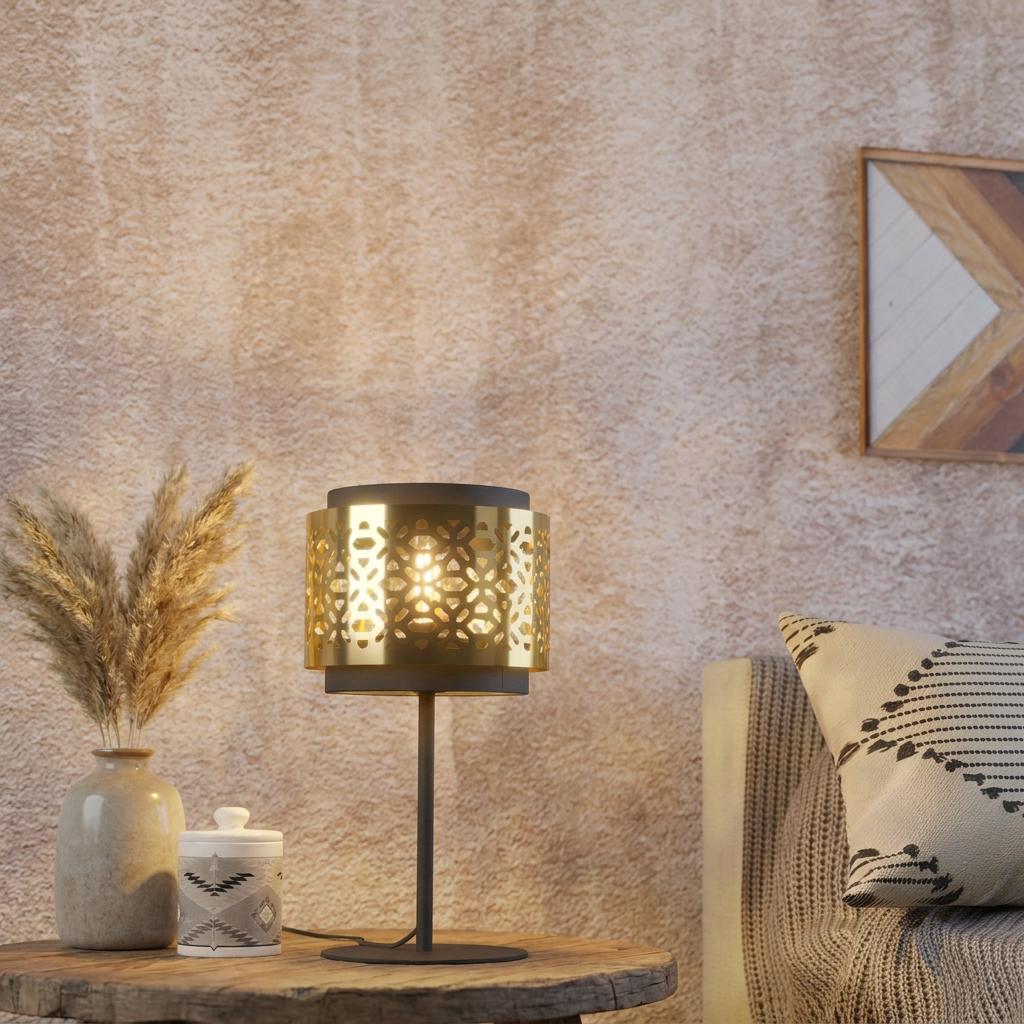 Lit black table lamp with a perforated brass shade on a wooden table beside a vase of pampas grass.