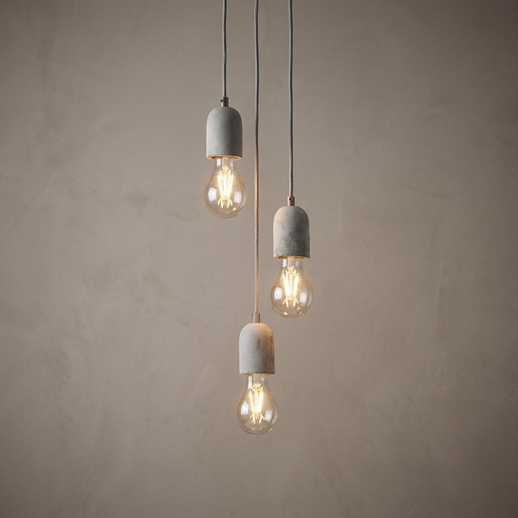Industrial triple pendant light with concrete holders and filament bulbs hanging against a textured neutral wall.