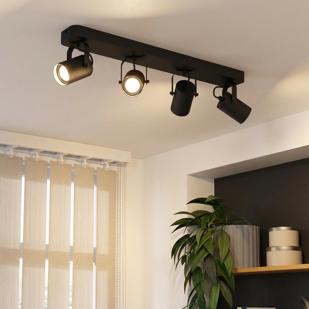 Modern black four-light bar spotlight mounted on a white ceiling in a room with a plant and window blinds.