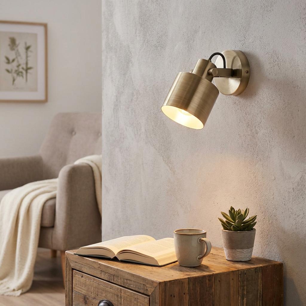 Brushed gold wall spotlight mounted on a textured wall, illuminating a book and mug on a wooden side table.