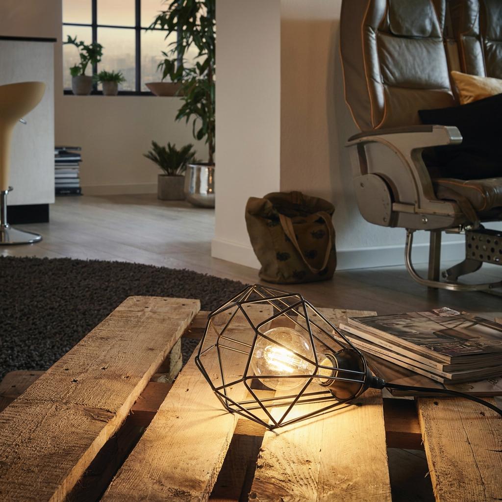A black geometric wire cage table lamp sits lit on a wooden pallet table in a modern industrial living room.