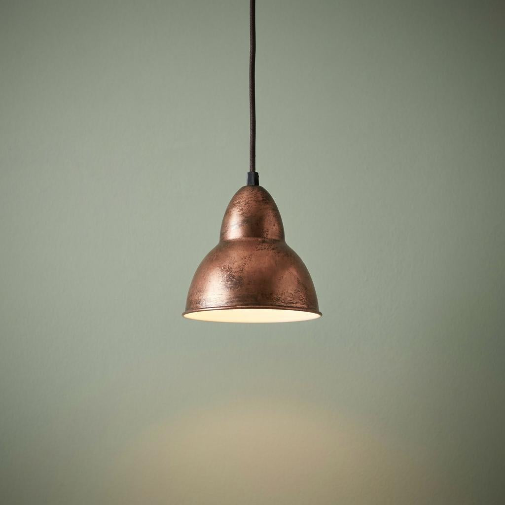 Distressed copper conical pendant light hanging against a sage green wall, emitting a warm glow.