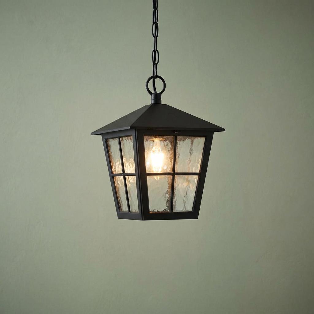 A black lantern-style outdoor pendant light with textured glass panels and a glowing bulb against a sage green wall.