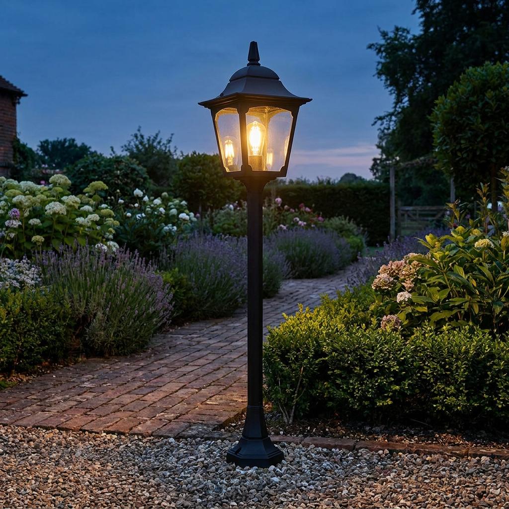 A black outdoor lantern bollard glowing at dusk on a brick path surrounded by garden flowers.
