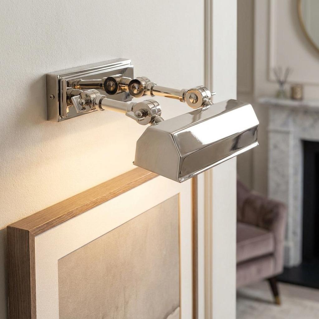 Polished nickel picture light with adjustable twin arms illuminating a framed artwork on a neutral wall.