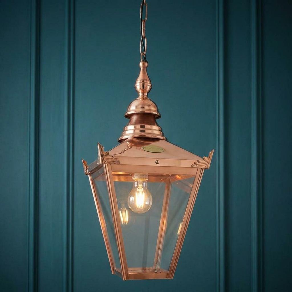 A polished copper lantern pendant light with clear glass panes hanging against a dark teal paneled background.