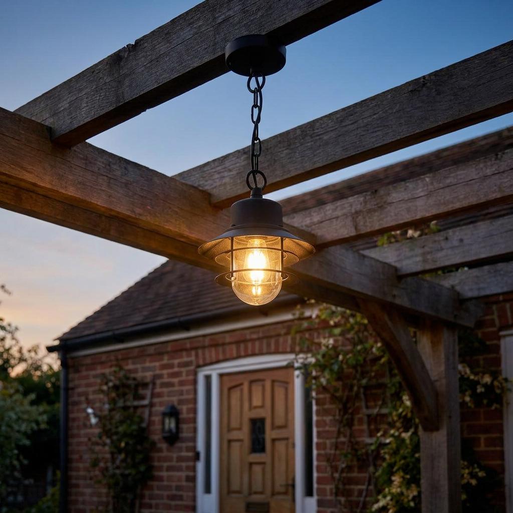 Black outdoor chain lantern hanging from a wooden pergola beam at dusk, casting a warm glow.